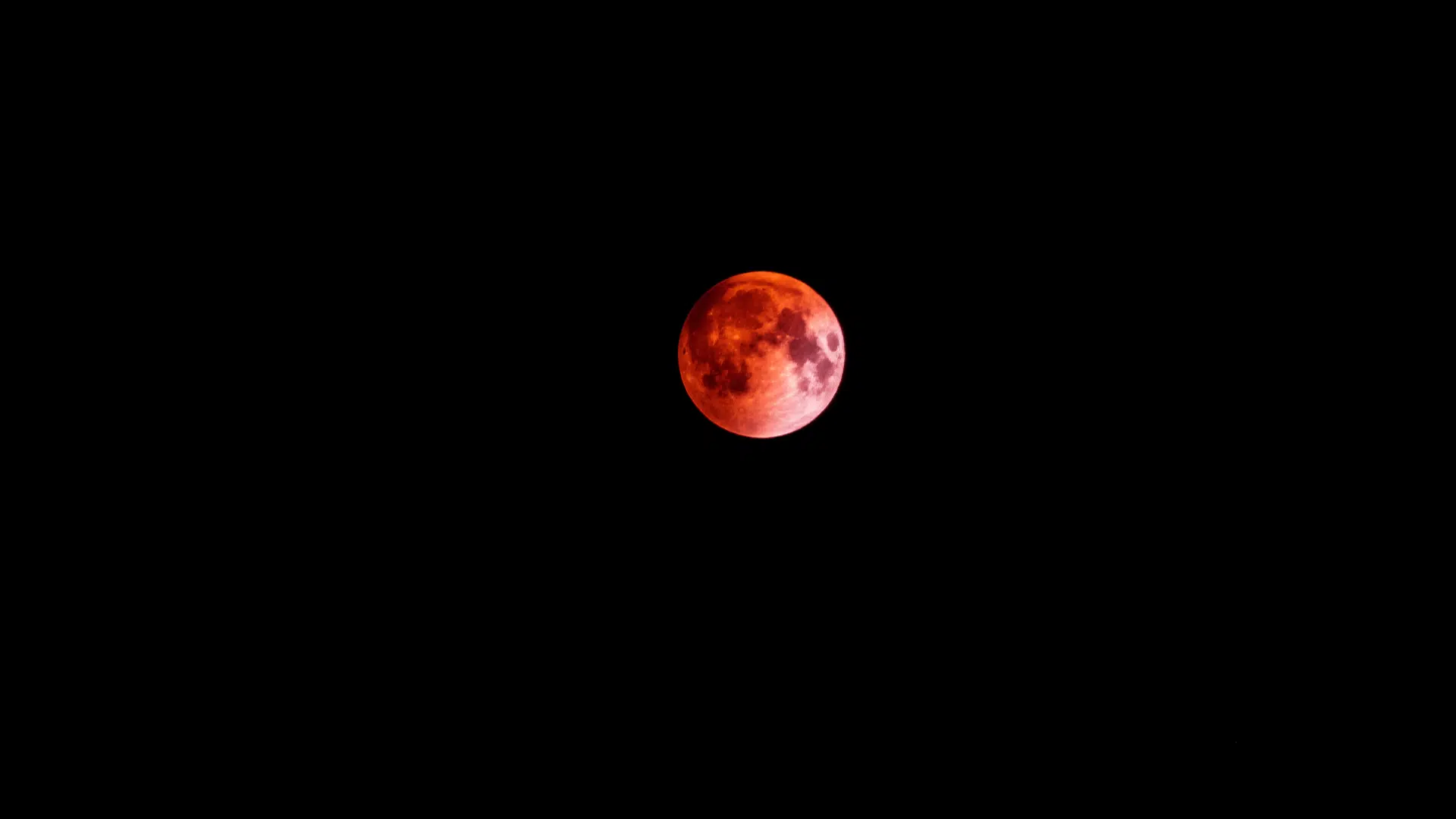 A red, shadowy lunar eclipse set against a black sky, with the moon appearing vivid and textured