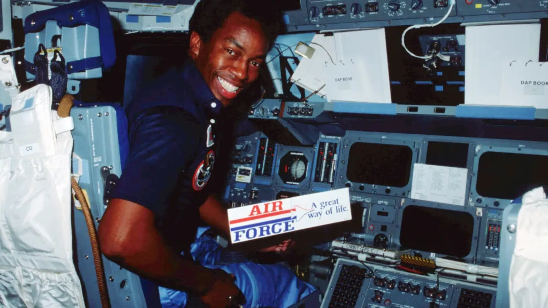 Astronaut Guion Bluford in a flight suit holding a sign inside the complex cockpit of a Space Shuttle