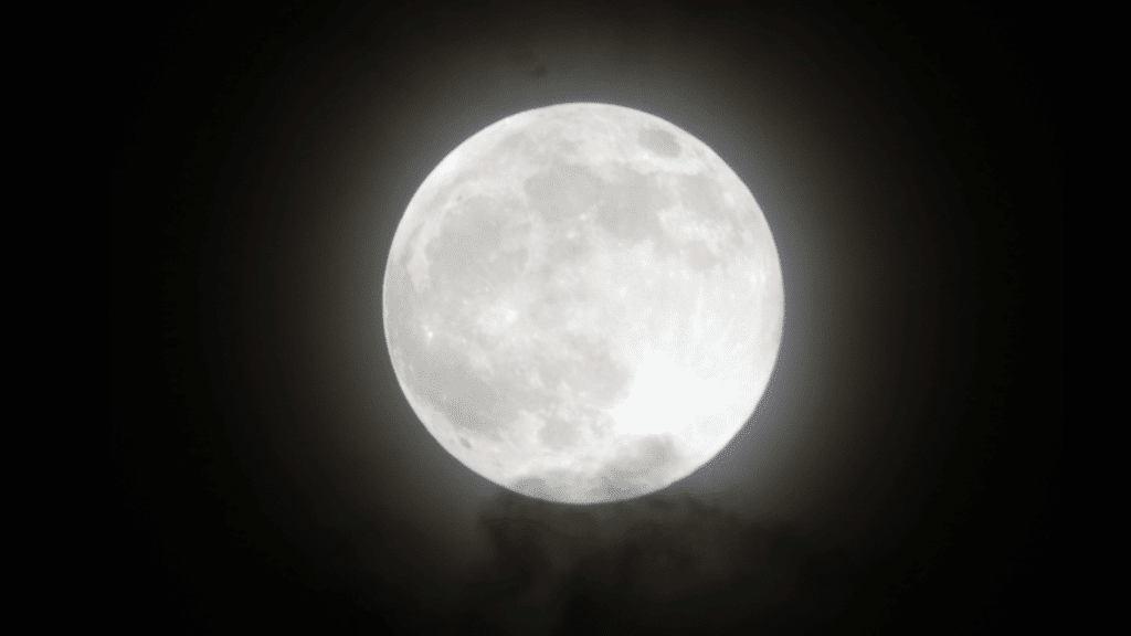 Bright, detailed full moon shining against a dark night sky, partially obscured by faint, wispy clouds below.