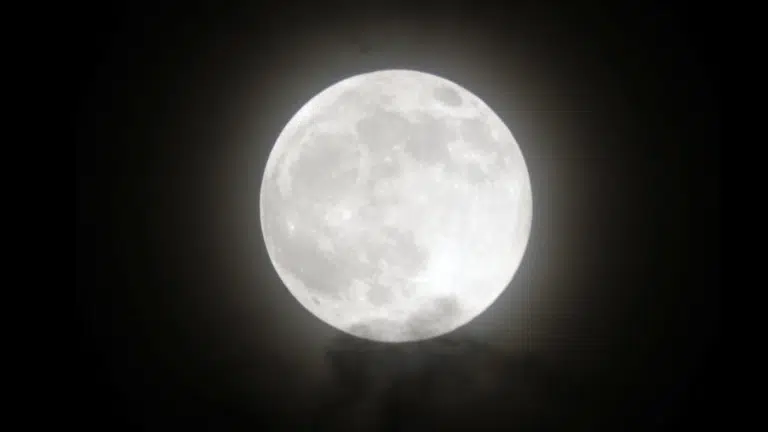 Bright, detailed full moon shining against a dark night sky, partially obscured by faint, wispy clouds below.