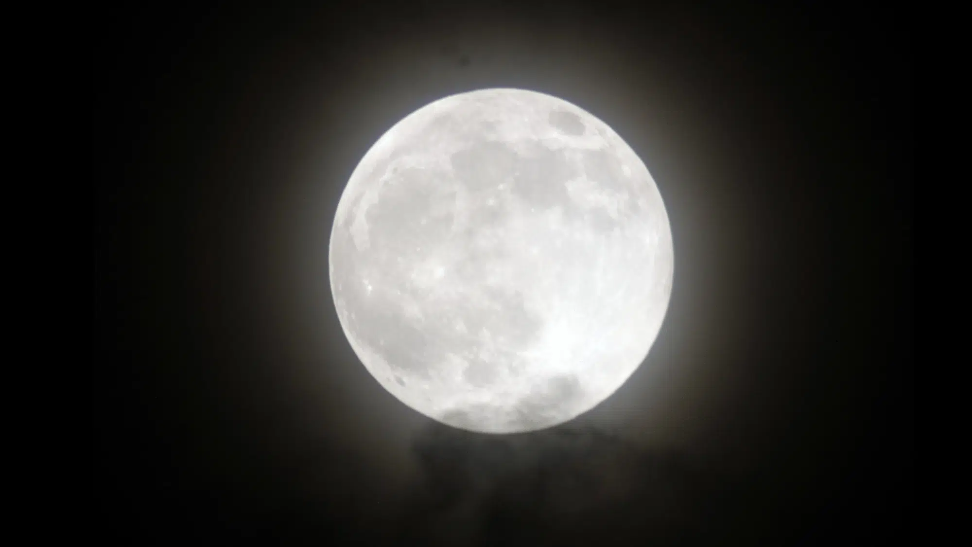 Bright, detailed full moon shining against a dark night sky, partially obscured by faint, wispy clouds below.