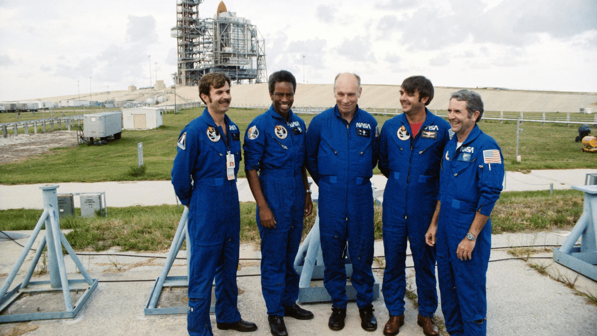 Five NASA astronauts in blue flight suits stand outdoors in front of a Space Shuttle on a launch tower under a cloudy sky