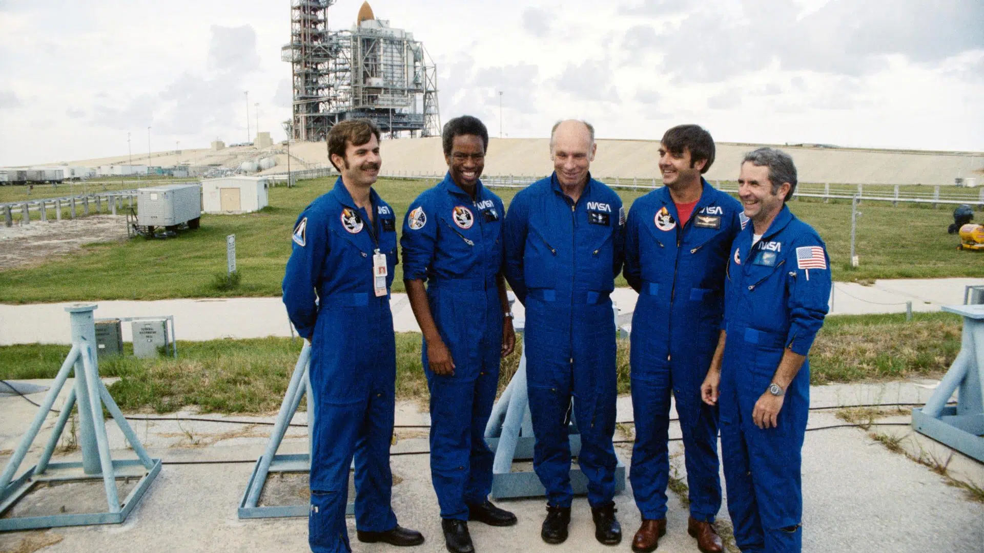 Five NASA astronauts in blue flight suits stand outdoors in front of a Space Shuttle on a launch tower under a cloudy sky