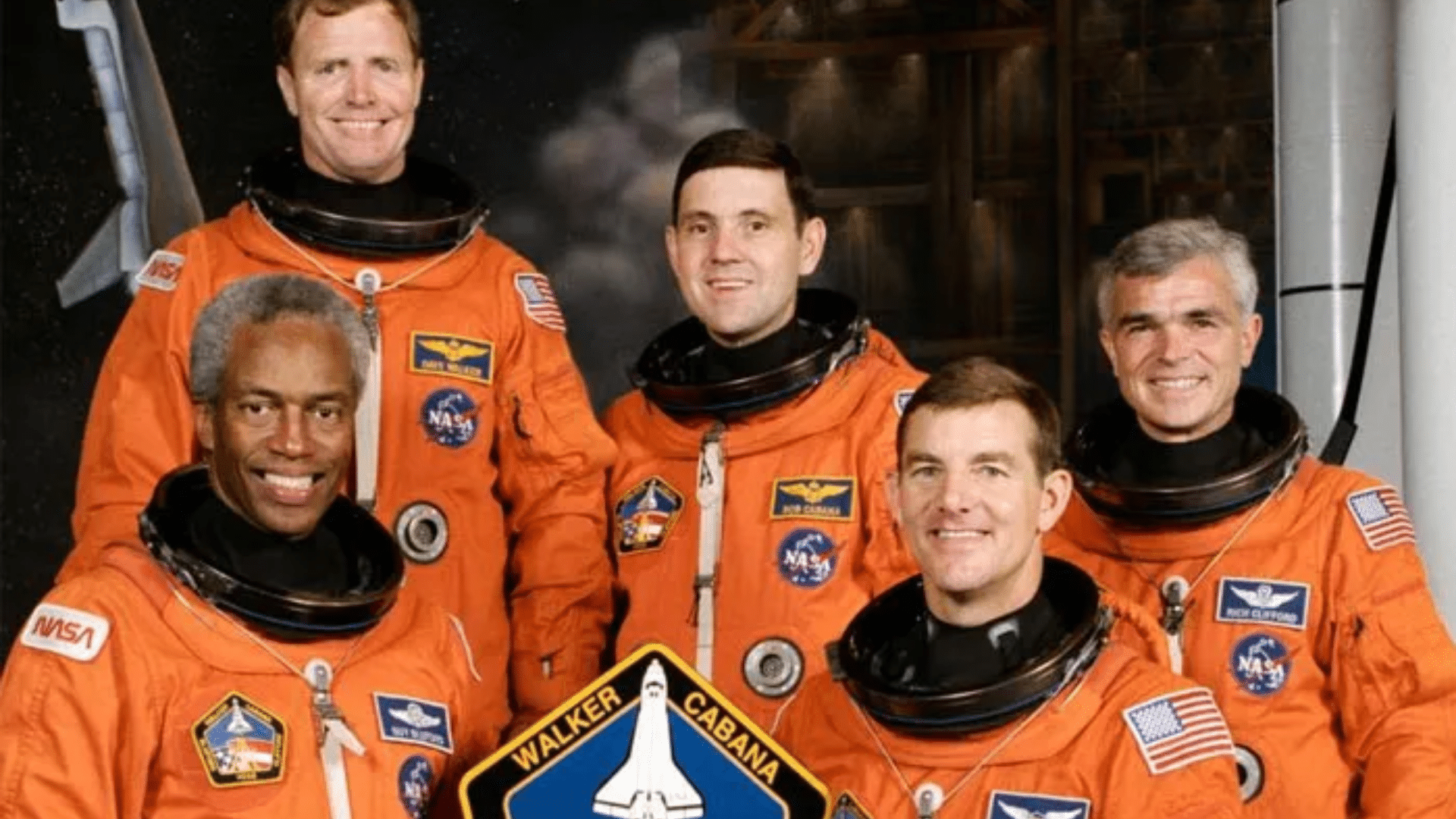 Five smiling NASA astronauts in orange launch suits pose with their mission patch, featuring a space shuttle launch silhouette
