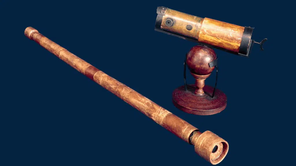 Galileo galilei telescope lies diagonally next to a smaller, brass and wood refracting telescope mounted on a dark wooden spherical stand against a blue background.