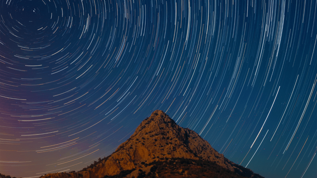 Long-exposure image of star trails curving across a deep blue night sky above a rugged, lit mountain peak