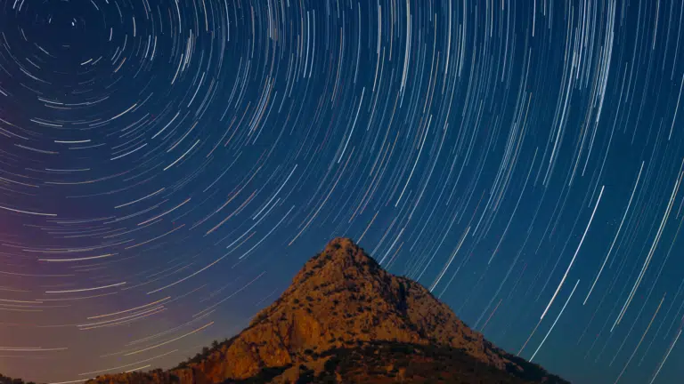 Long-exposure image of star trails curving across a deep blue night sky above a rugged, lit mountain peak