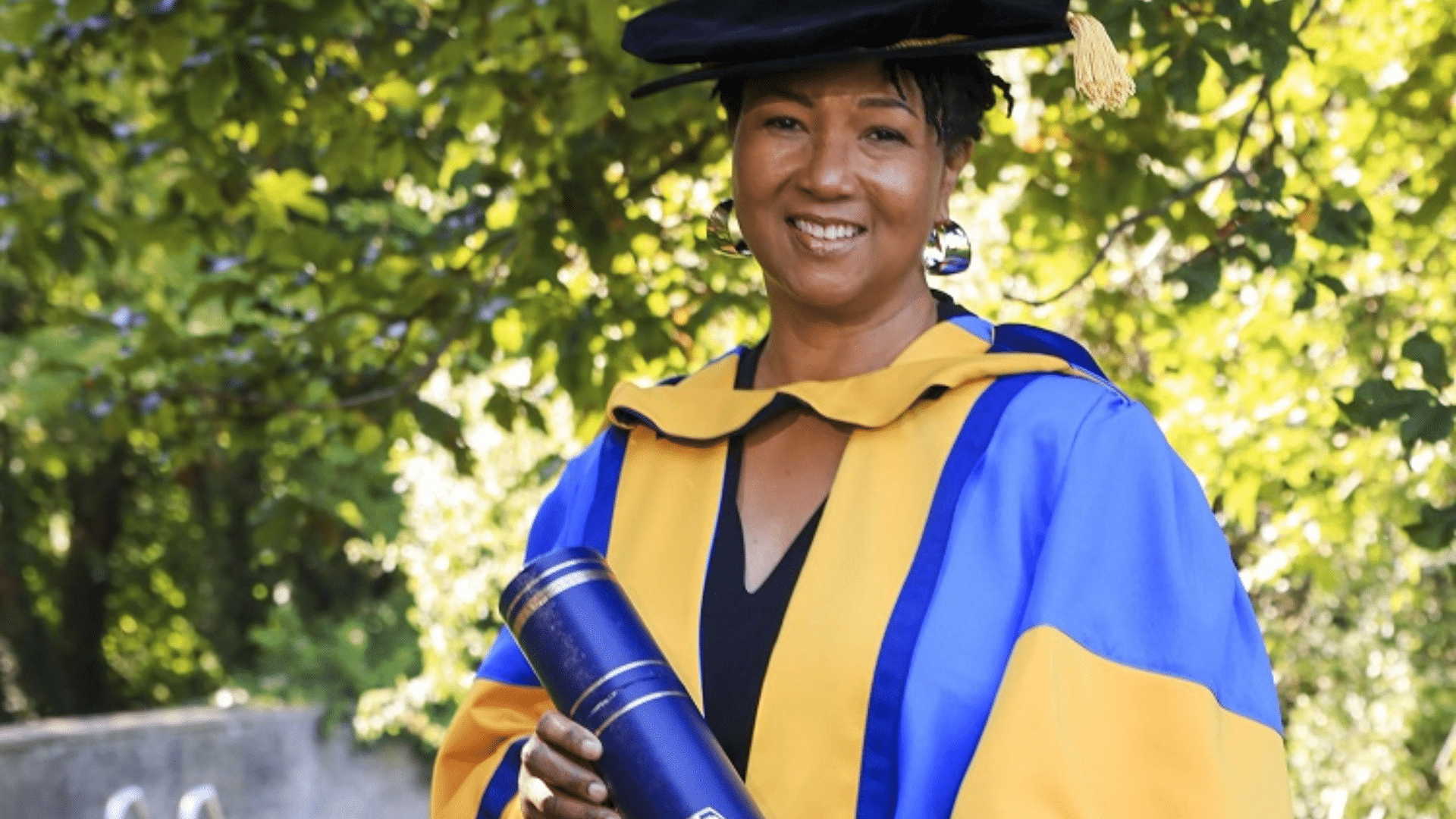 Mae Jemison in blue and yellow academic regalia and mortarboard holds a rolled diploma