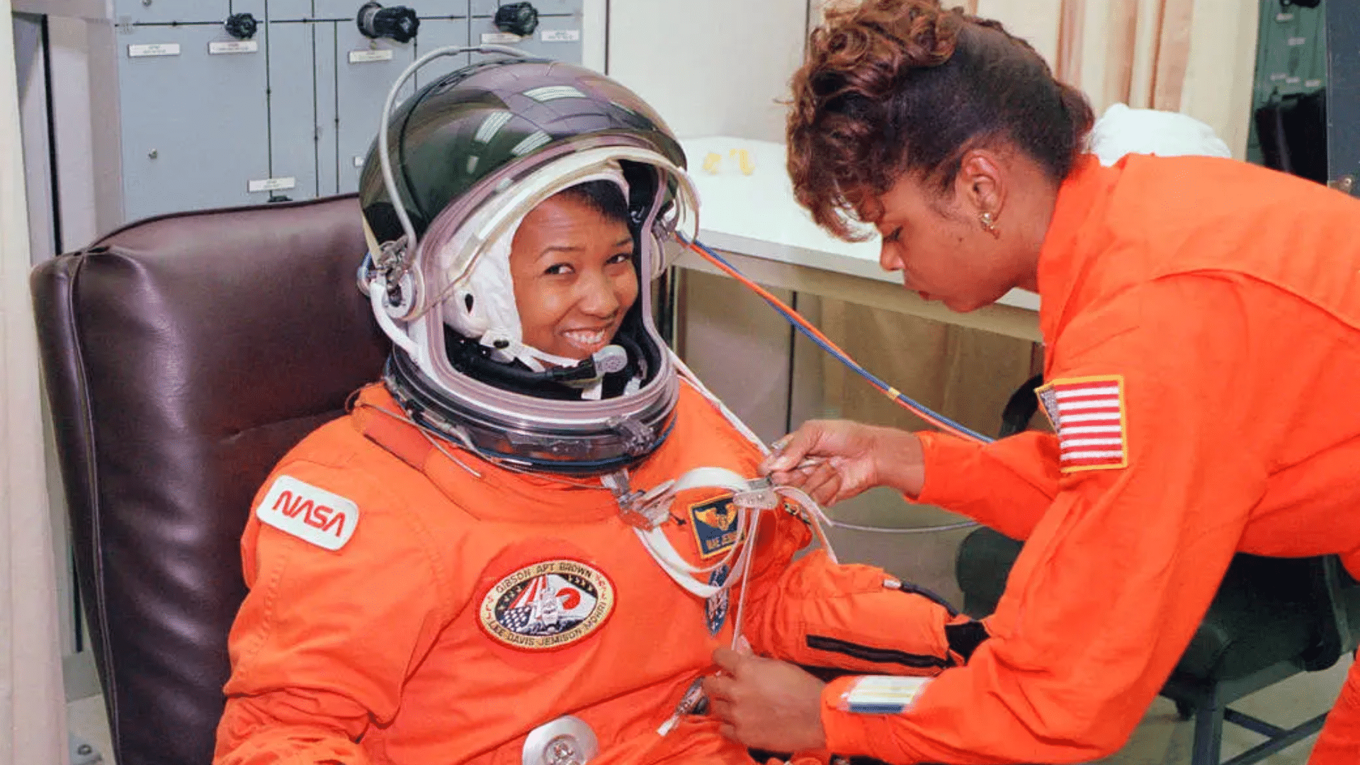 Mae Jemison in orange flight suit, smiling wearing a space helmet, undergo suit fitting with tubing connections.
