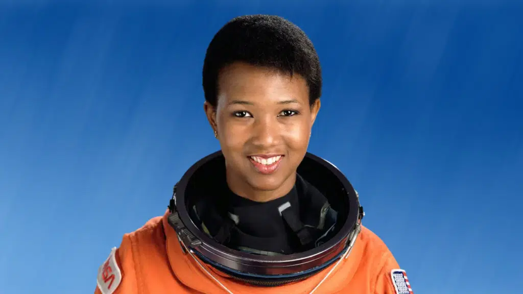 Portrait of astronaut Mae Jemison smiling in an orange flight suit against a bright blue background.