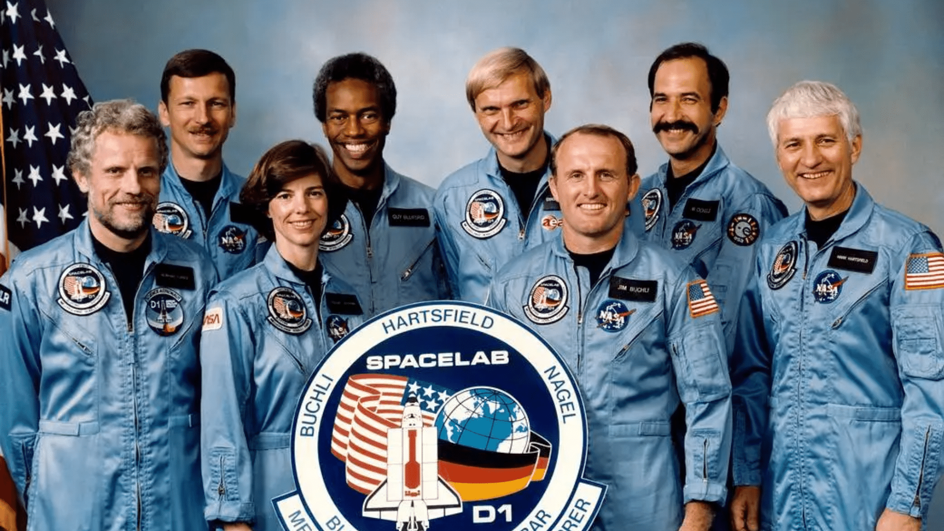 Seven astronauts in light blue flight suits pose for a group portrait next to an American flag while holding the circular Spacelab D-1 mission patch