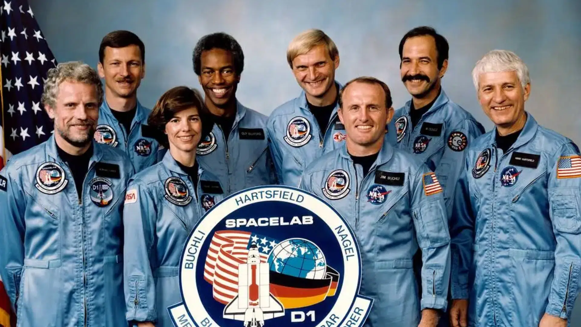 Seven astronauts in light blue flight suits pose for a group portrait next to an American flag while holding the circular Spacelab D-1 mission patch