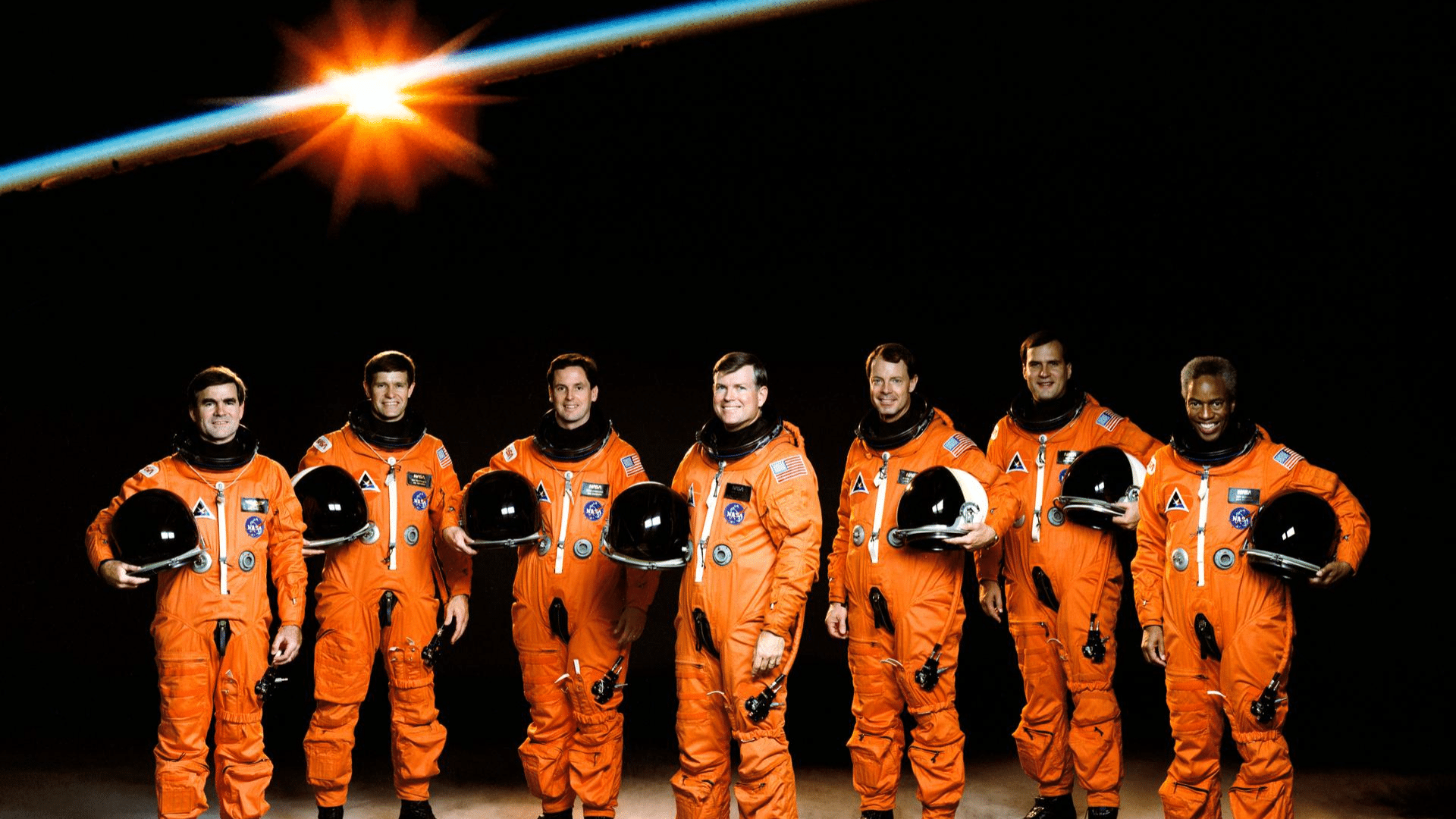 Seven astronauts in orange launch and entry suits stand against a dark background