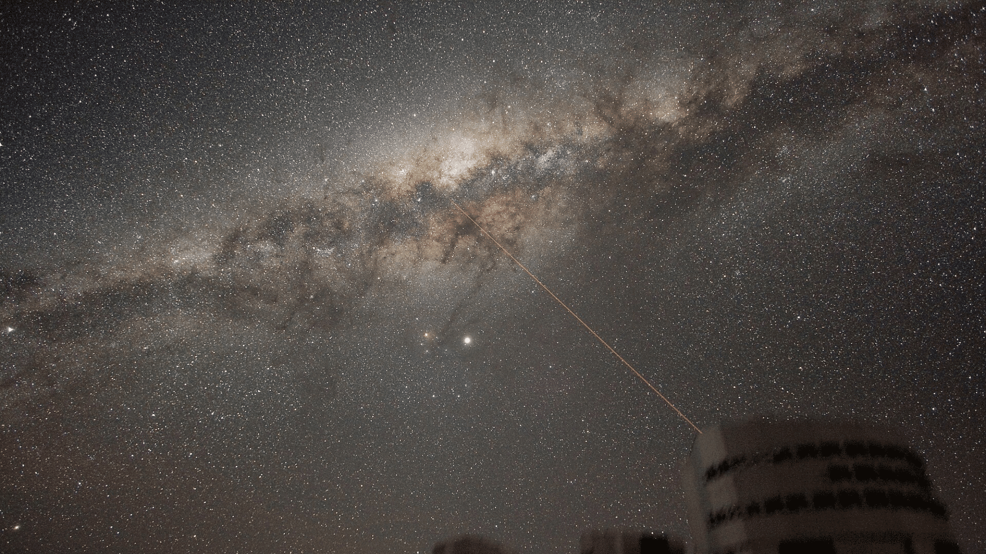 Telescope domes under a dense, star-filled sky featuring the bright band of the Milky Way and a distinct red laser beam pointing upward.