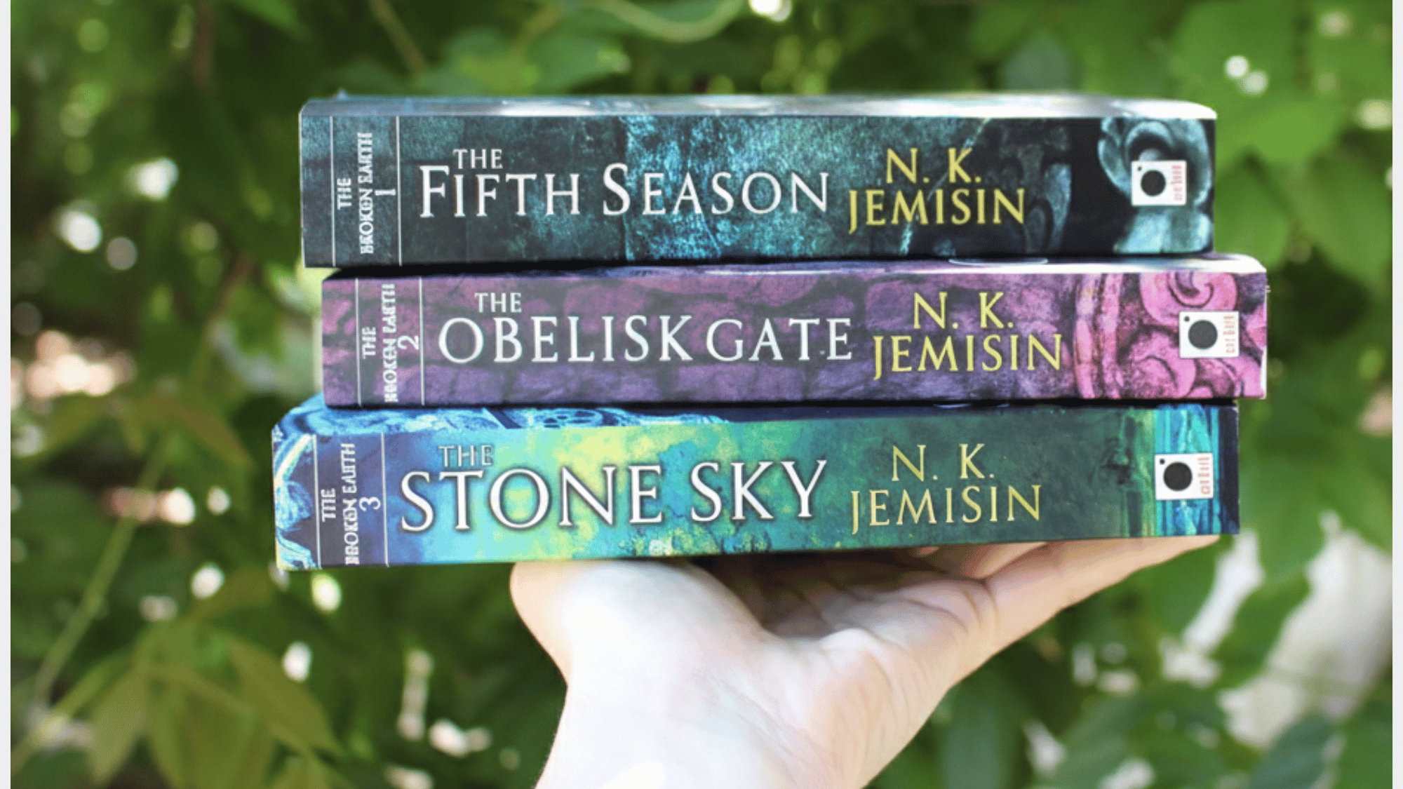 Three paperback books from N.K. Jemisin's Broken Earth trilogy The Fifth Season, The Obelisk Gate, and The Stone Sky are held stacked in a hand against a blurred green outdoor background.