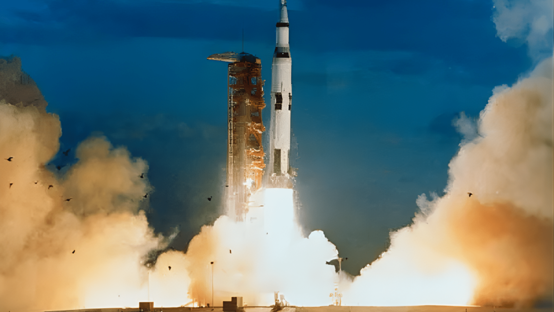 A Saturn V rocket launches from a launch complex over water, kicking up massive plumes of smoke and fire