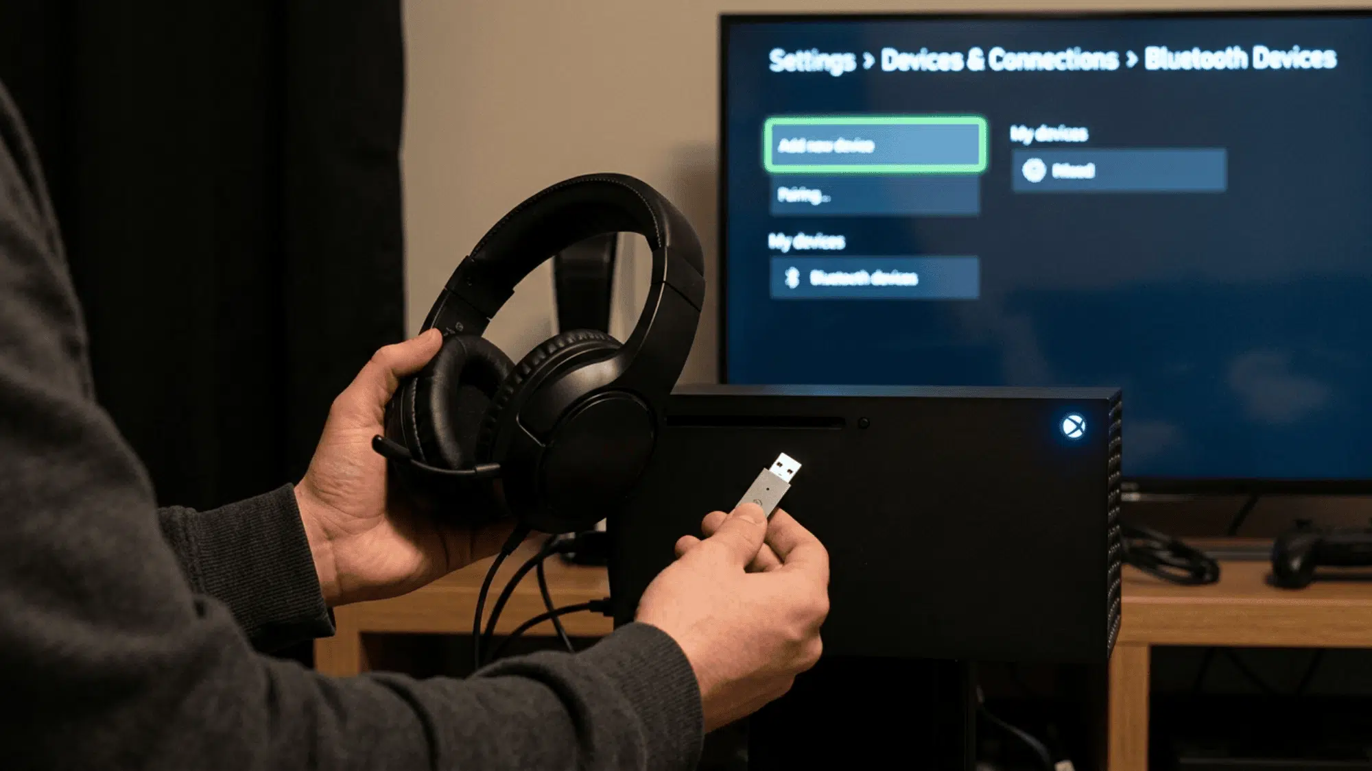 Person plugging USB Bluetooth adapter into Xbox while holding headphones near screen