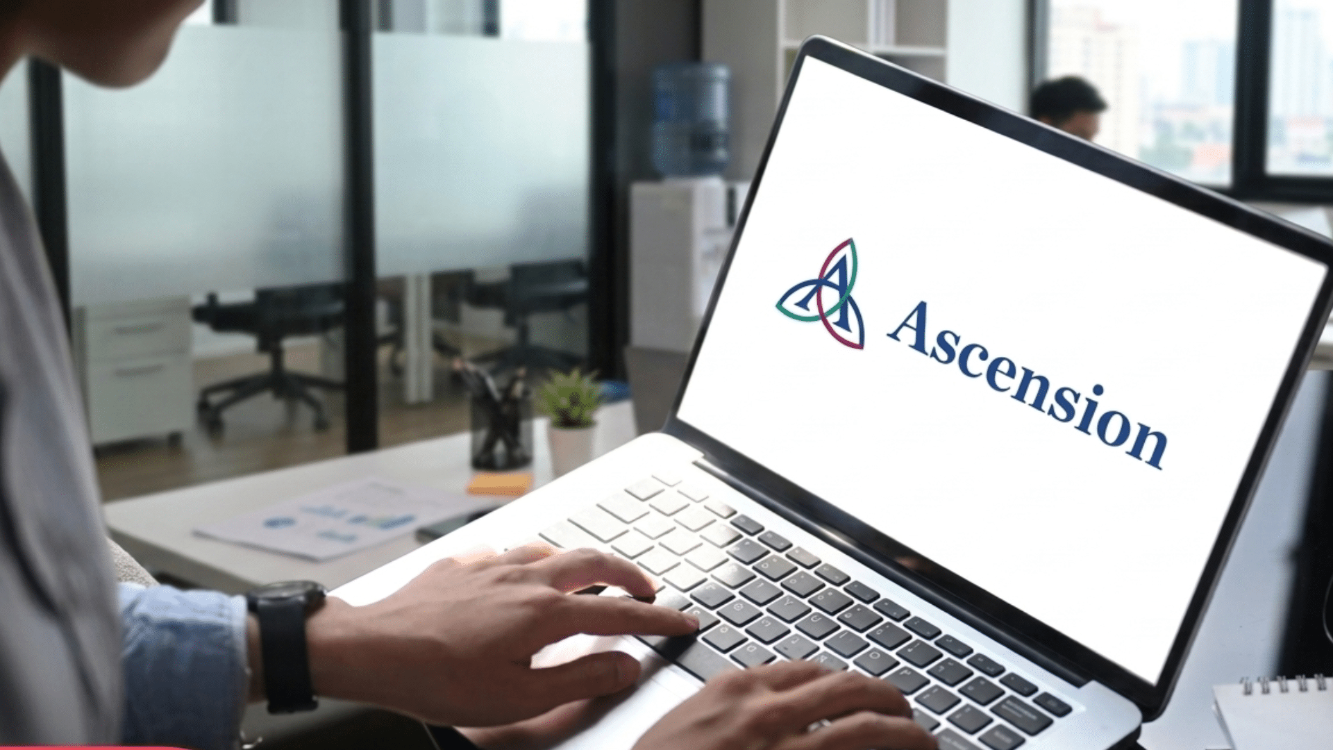 Person typing on a laptop displaying the Ascension logo in a modern office setting