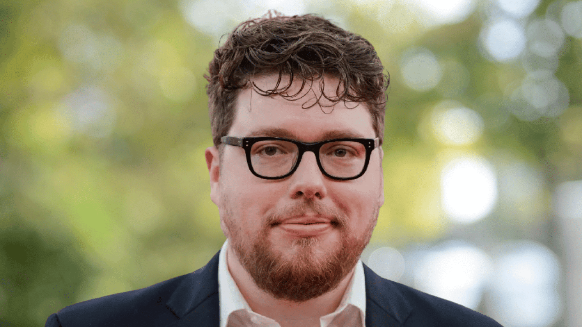 Michael Sarnoski director wearing black glasses and navy blazer with blurred green background