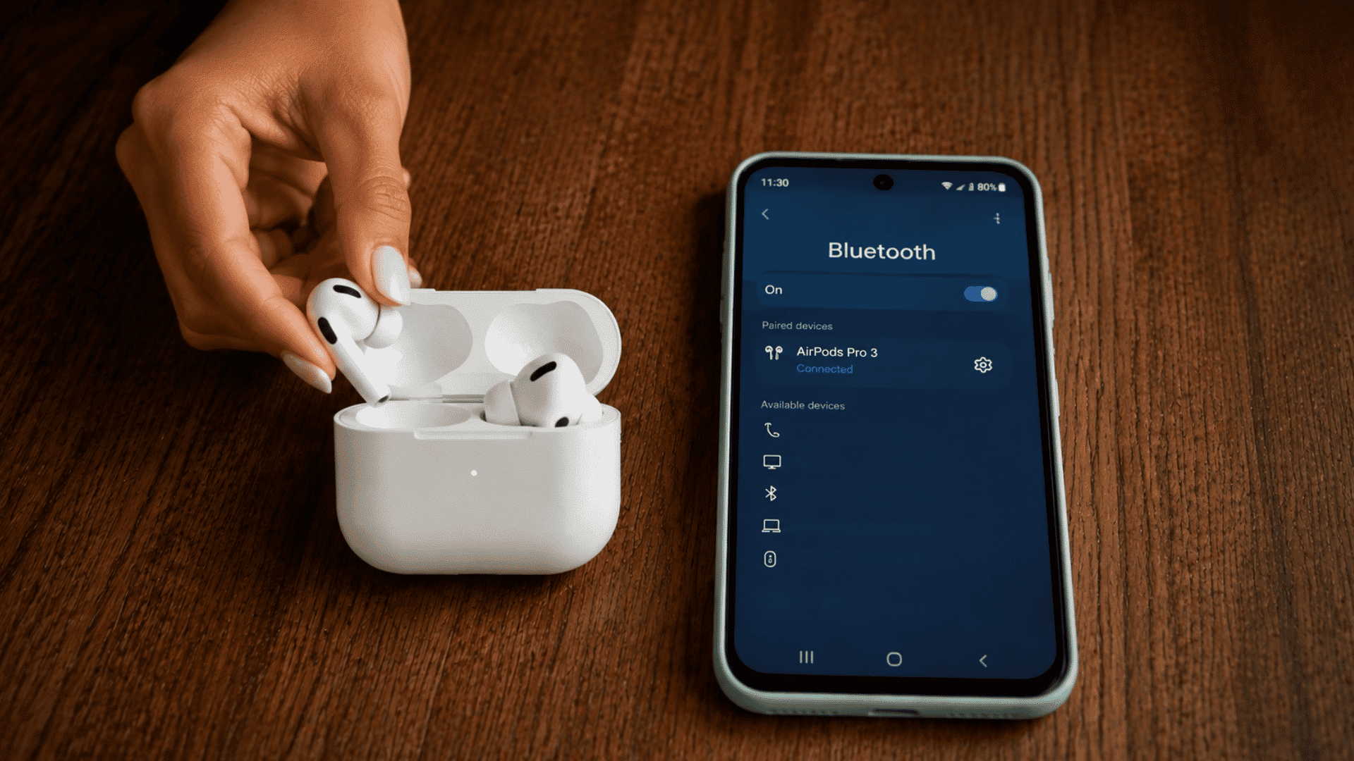 airPods Pro connected to an Android phone via Bluetooth, with one earbud being removed from the charging case on a wooden desk.