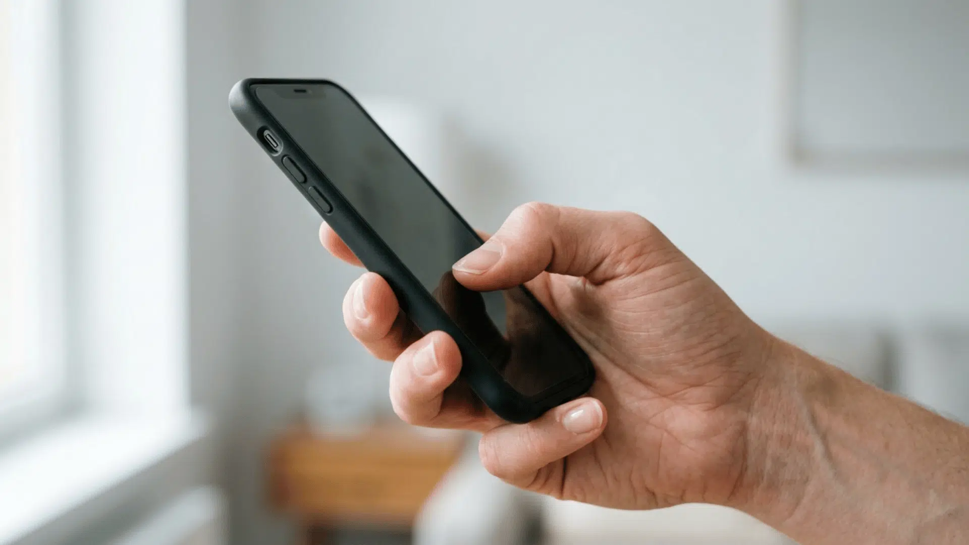 hand holding a black smartphone in a bright indoor setting with soft natural daylight and a blurred light background